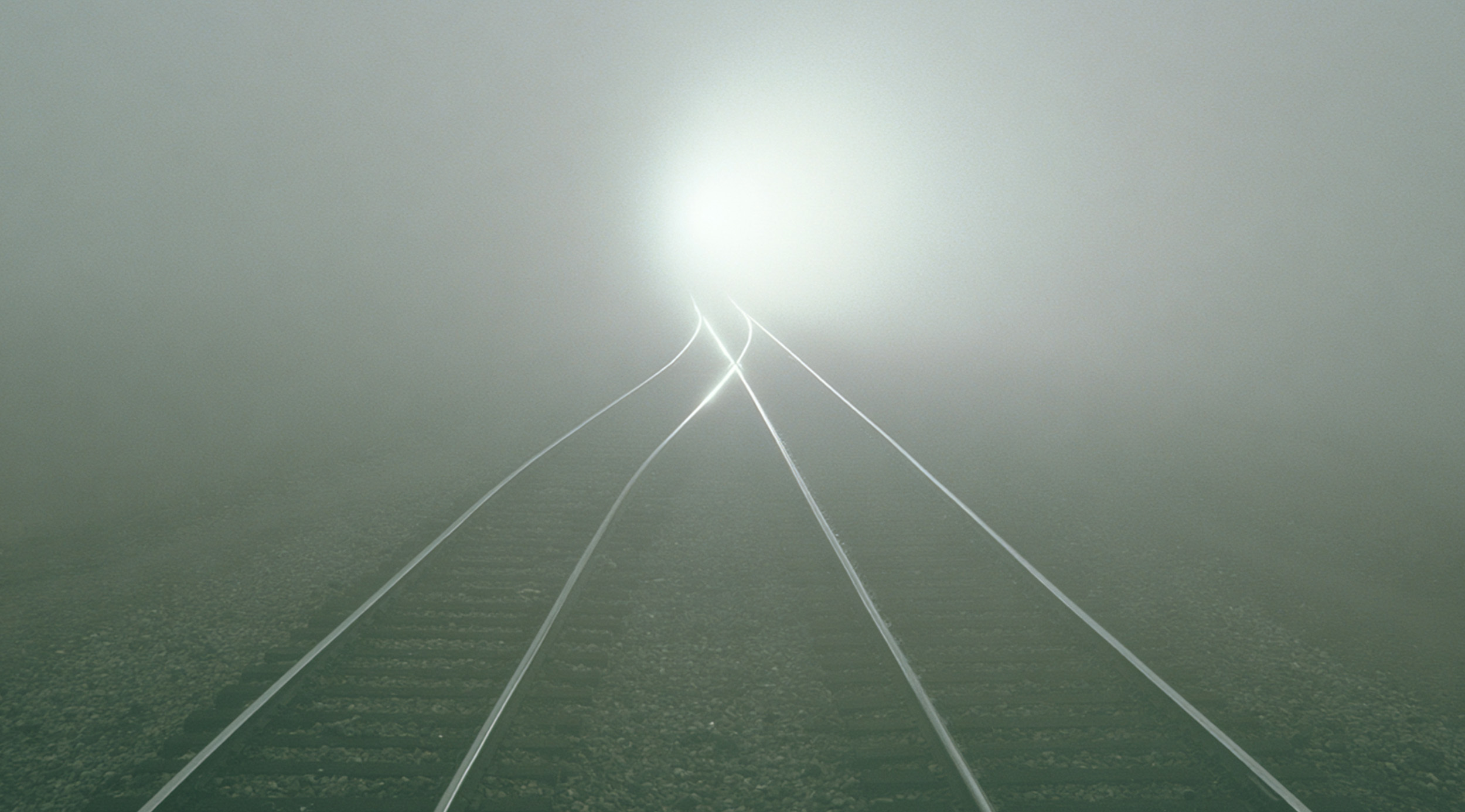 Foggy railway tracks splitting subtly into diverging paths before disappearing into a bright white haze, creating a cinematic metaphor for systems drifting off-course while continuing forward.