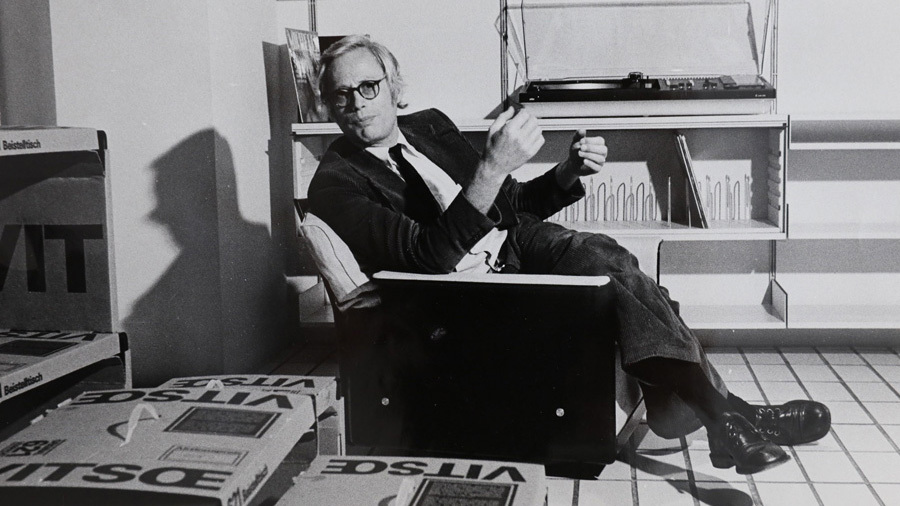 Dieter Rams seated in a chair surrounded by Vitsoe packaging and modernist furniture.